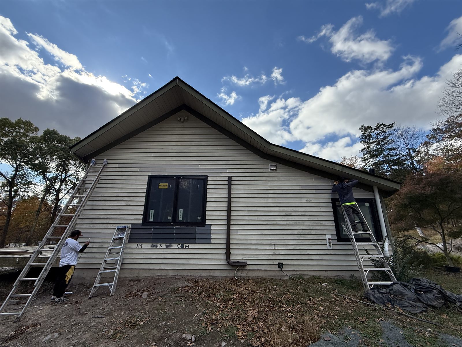 Gable end of a house being prepared for painting, with two painters on ladders and weathered white siding