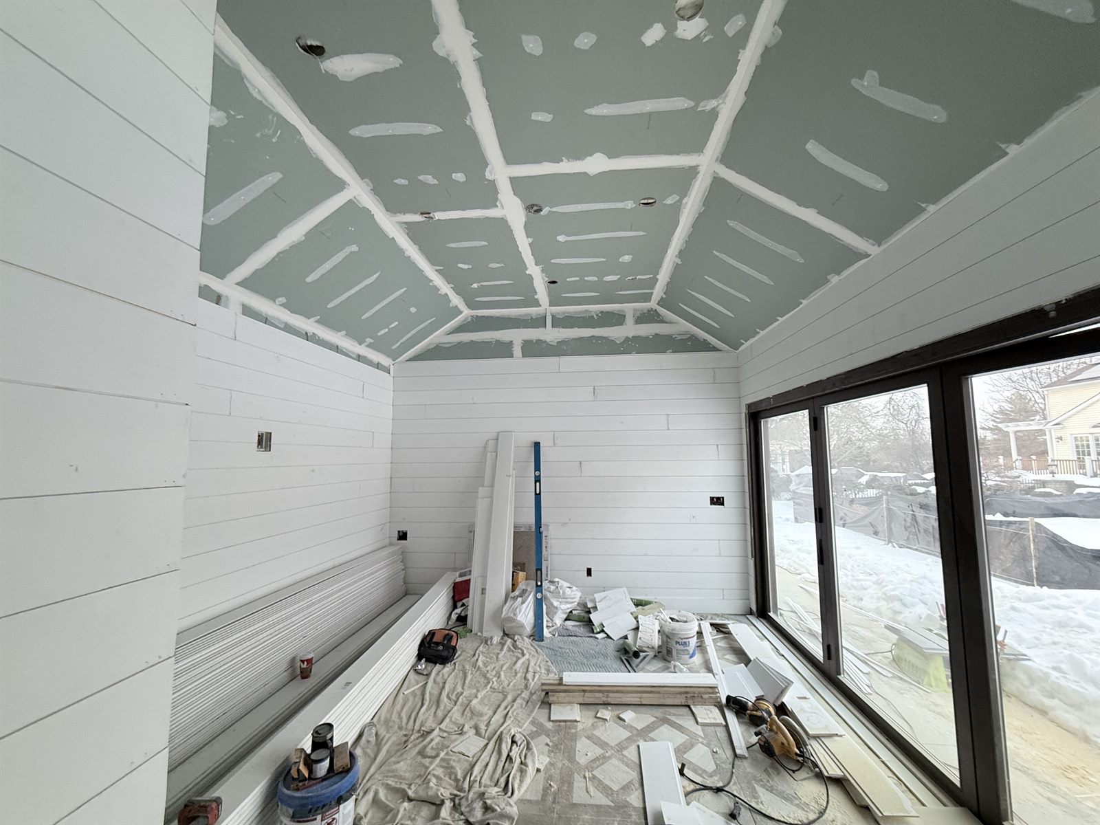 Sunroom under construction with unfinished drywall ceiling, taped seams, primed shiplap walls and tools on the floor